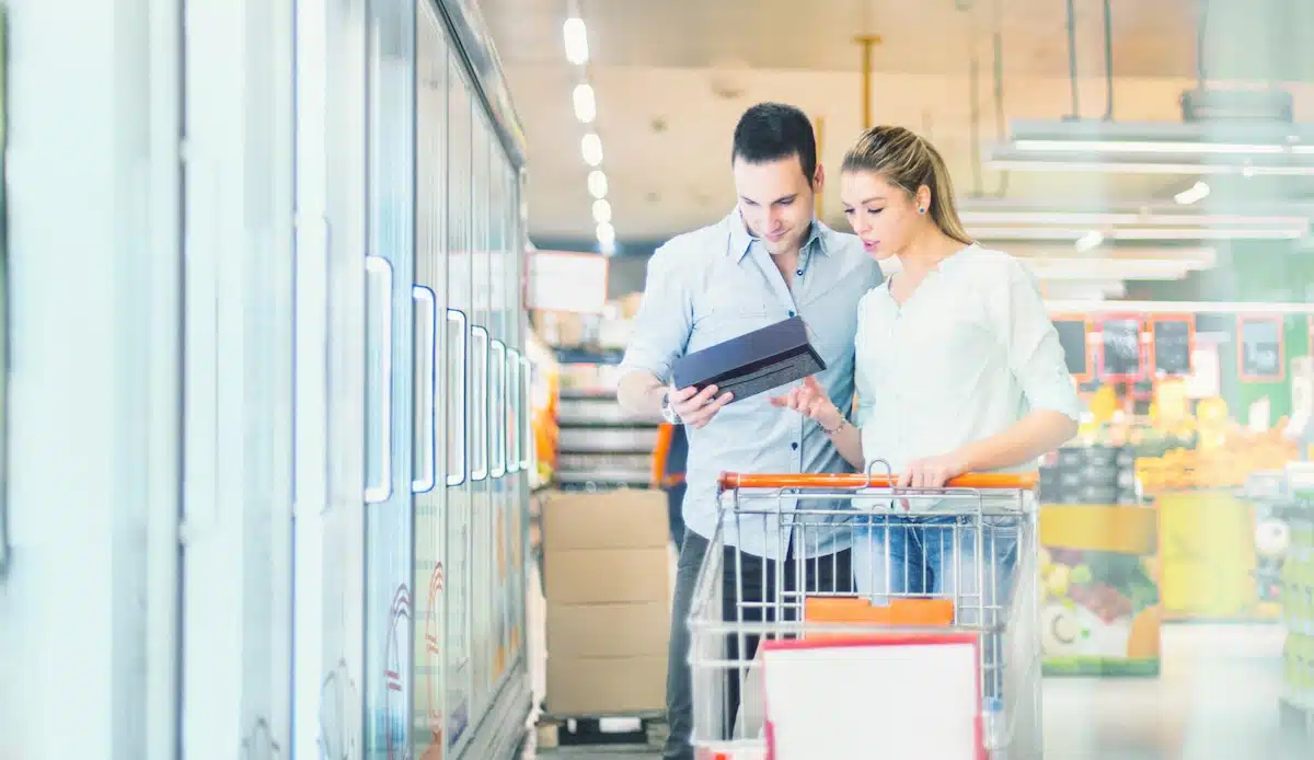 Junges erwachsenes Paar kauft Tiefkühlkost im örtlichen Supermarkt. Sie stehen neben der Gefrierabteilung und lesen Etiketten und Preisschilder.