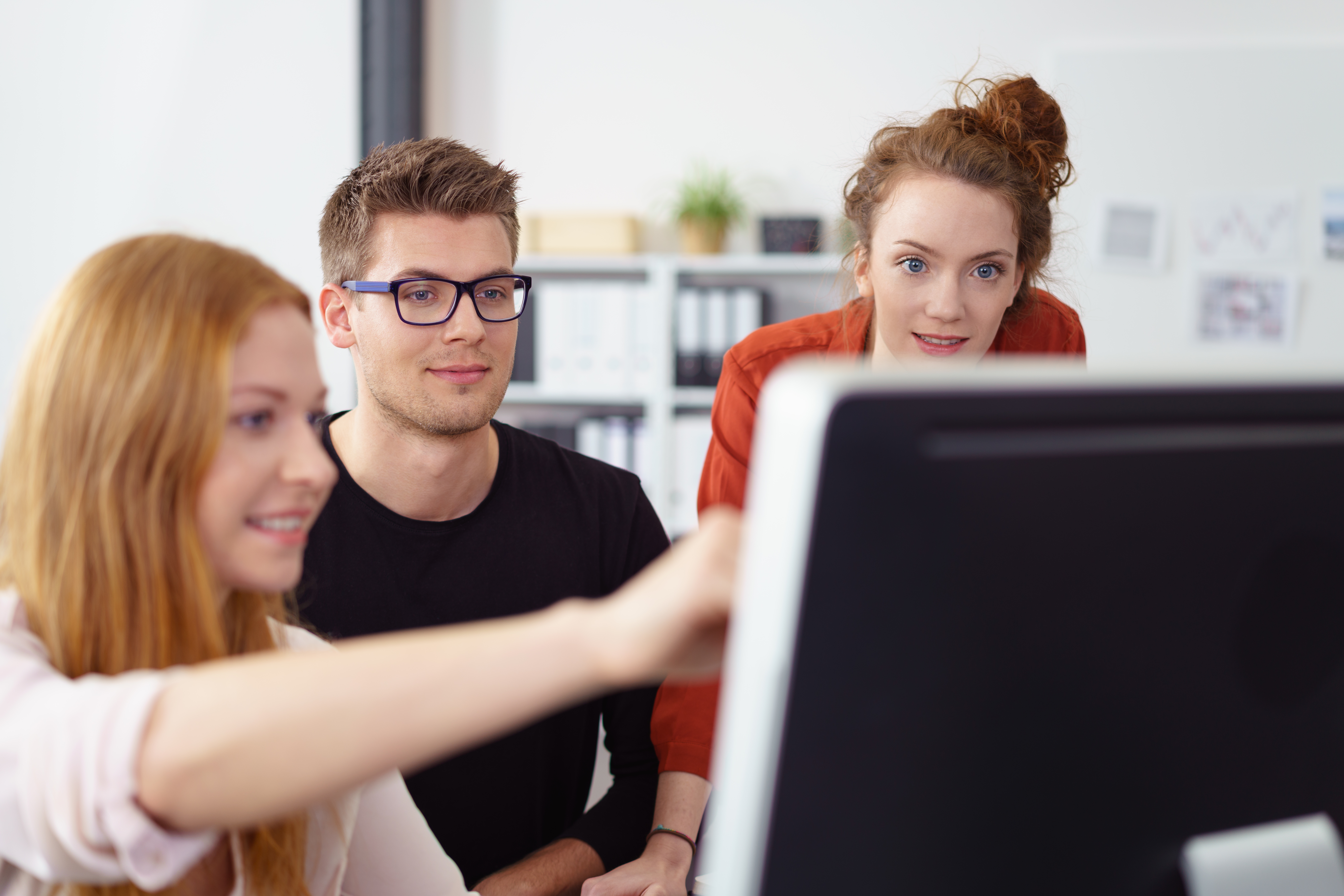 Junges motiviertes Business-Team aus zwei Frauen und einem jungen Mann, die zusammen an einem Desktop-Computer arbeiten, um Informationen auf dem Bildschirm zu diskutieren, konzentrieren Sie sich auf den jungen Mann