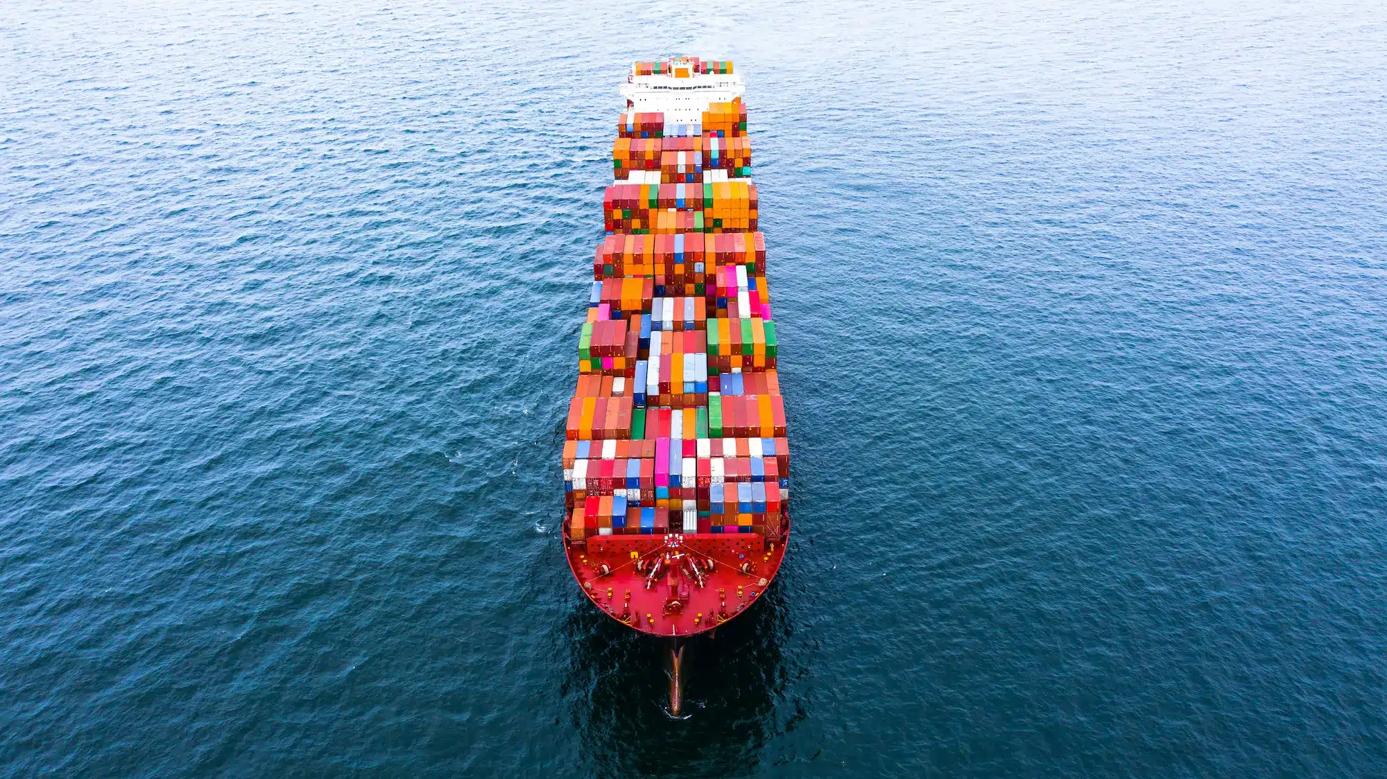Luftaufnahme eines großen Containerschiffs, das mit bunten Frachtcontainern beladen über dunkelblaues Meer fährt.